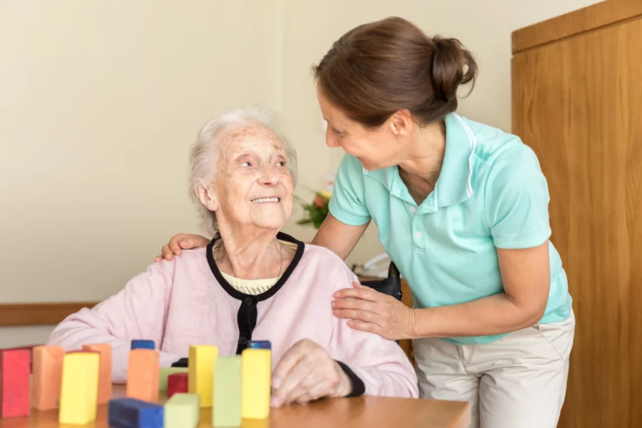 Pflegerin beugt sich zu einer älteren Frau, vor ihr auf dem Tisch stehen therapeutische Beschäftigungsklötze