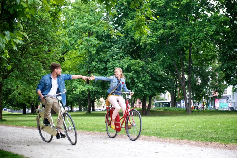 Junger Mann und junge Frau auf Fahrrädern , im Hintergrund Wiese und Bäume sowie Gebäude